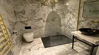 Luxurious marble-tiled bathroom in Manchester featuring a black walk-in shower tray, gold fixtures, wall-hung toilet, and a sleek vanity with a black basin.