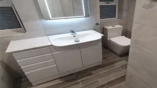 Modern bathroom in Alderley Edge featuring a sleek white vanity with integrated sink, LED mirror, and a contemporary toilet on wood-look flooring.